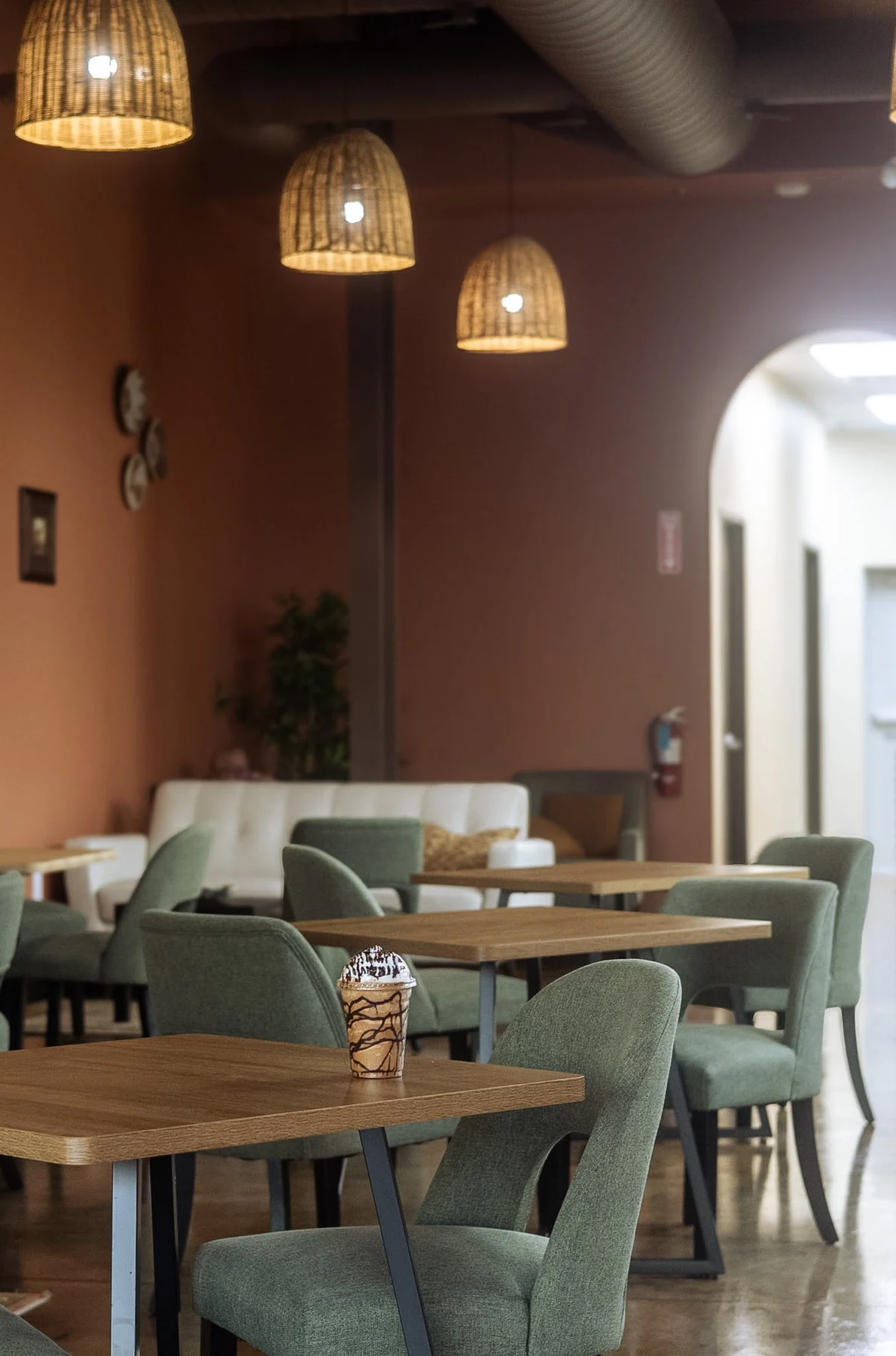 Elura’s sunlit lounge with sage green chairs, woven pendant lights, and a chocolate drink on a café table