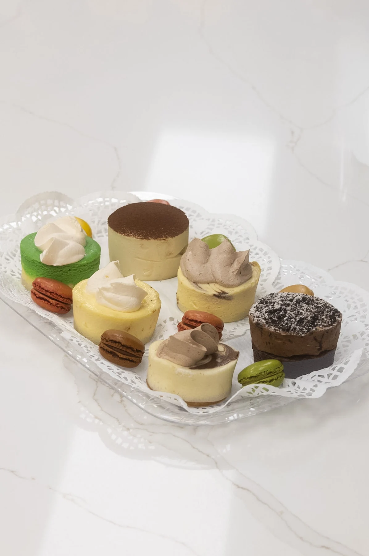 A selection of petite cakes with cream toppings, chocolate, and colorful macarons on a glass platter